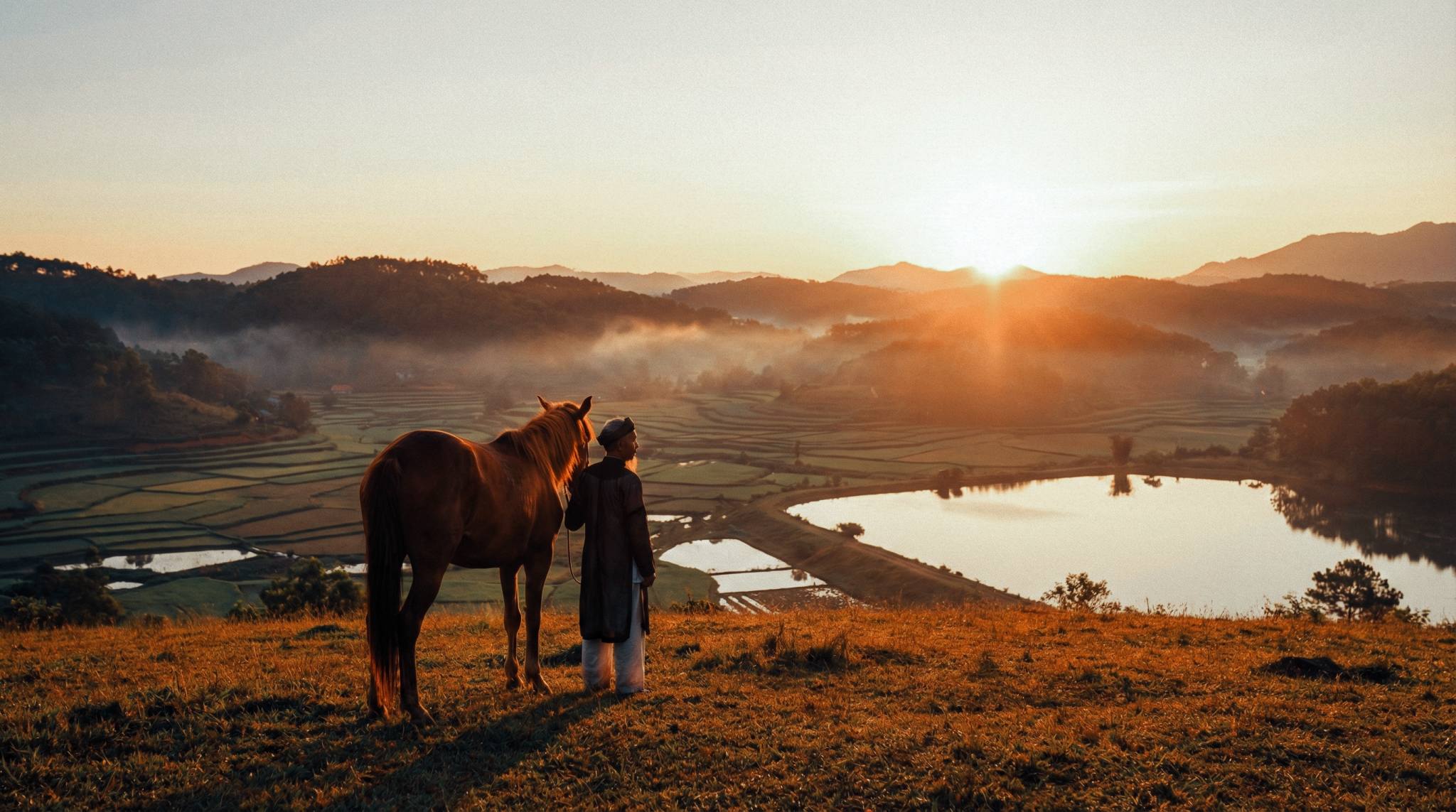 Cảnh bình minh vùng cao Việt Nam: Người đàn ông đứng lặng yên bên con ngựa không yên cương, nhìn xuống thung lũng và hồ nước yên bình.